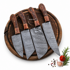 Four Damascus steel kitchen knives with patterned steel blades and reddish-brown wooden handles, arranged on a round wooden cutting board with small food props in the corner.