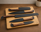 Five kitchen knives with dark textured blades and brown wooden handles are arranged on a light wood cutting board. The board sits on a wooden countertop with a tiled backsplash and a small container visible in the background.