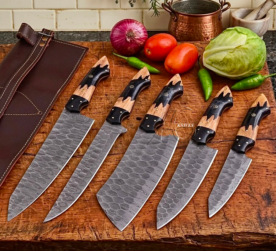 Five kitchen knives with patterned steel blades and wood-and-black handles are arranged on a wooden cutting board beside a brown leather sheath. Fresh vegetables including tomatoes, cabbage, green peppers, and an onion are placed behind the knives, with a copper pot and tiled wall in the background.