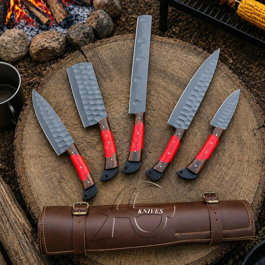 Five kitchen knives with textured silver blades and red handles are arranged on a round wooden slice, with a brown leather knife case in front. The background shows a rustic outdoor cooking scene with fire, rocks, and corn on the cob.