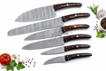 A set of seven Damascus steel kitchen knives with dark brown wooden handles and brass bolsters, arranged diagonally on a white background with culinary props like a tomato, garlic, peppercorns, and rosemary.