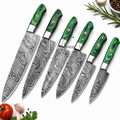 Six kitchen knives with patterned steel blades and green wood handles are arranged diagonally on a white background with small food props including a tomato, garlic, parsley, rosemary, and a bowl of peppercorns.