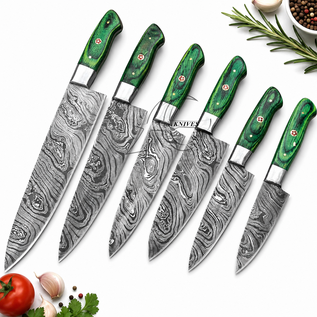 Six kitchen knives with patterned steel blades and green wood handles are arranged diagonally on a white background with small food props including a tomato, garlic, parsley, rosemary, and a bowl of peppercorns.