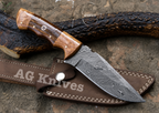 A fixed-blade knife with a patterned Damascus-style steel blade and a brown wood handle, resting on a brown leather sheath over a textured outdoor surface with a dark branch behind it. The sheath and blade show the AG Knives branding.