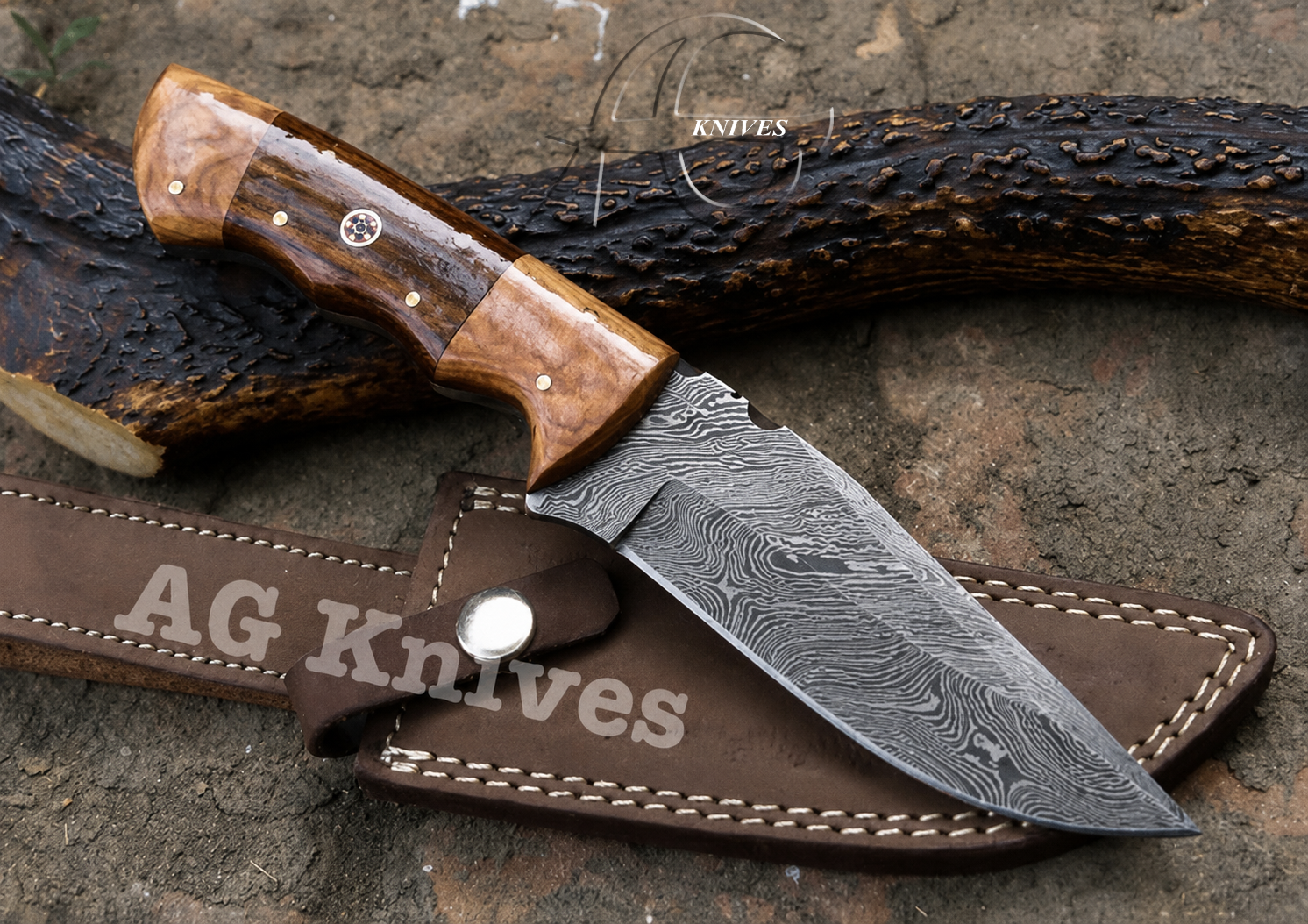 A fixed-blade knife with a patterned Damascus-style steel blade and a brown wood handle, resting on a brown leather sheath over a textured outdoor surface with a dark branch behind it. The sheath and blade show the AG Knives branding.