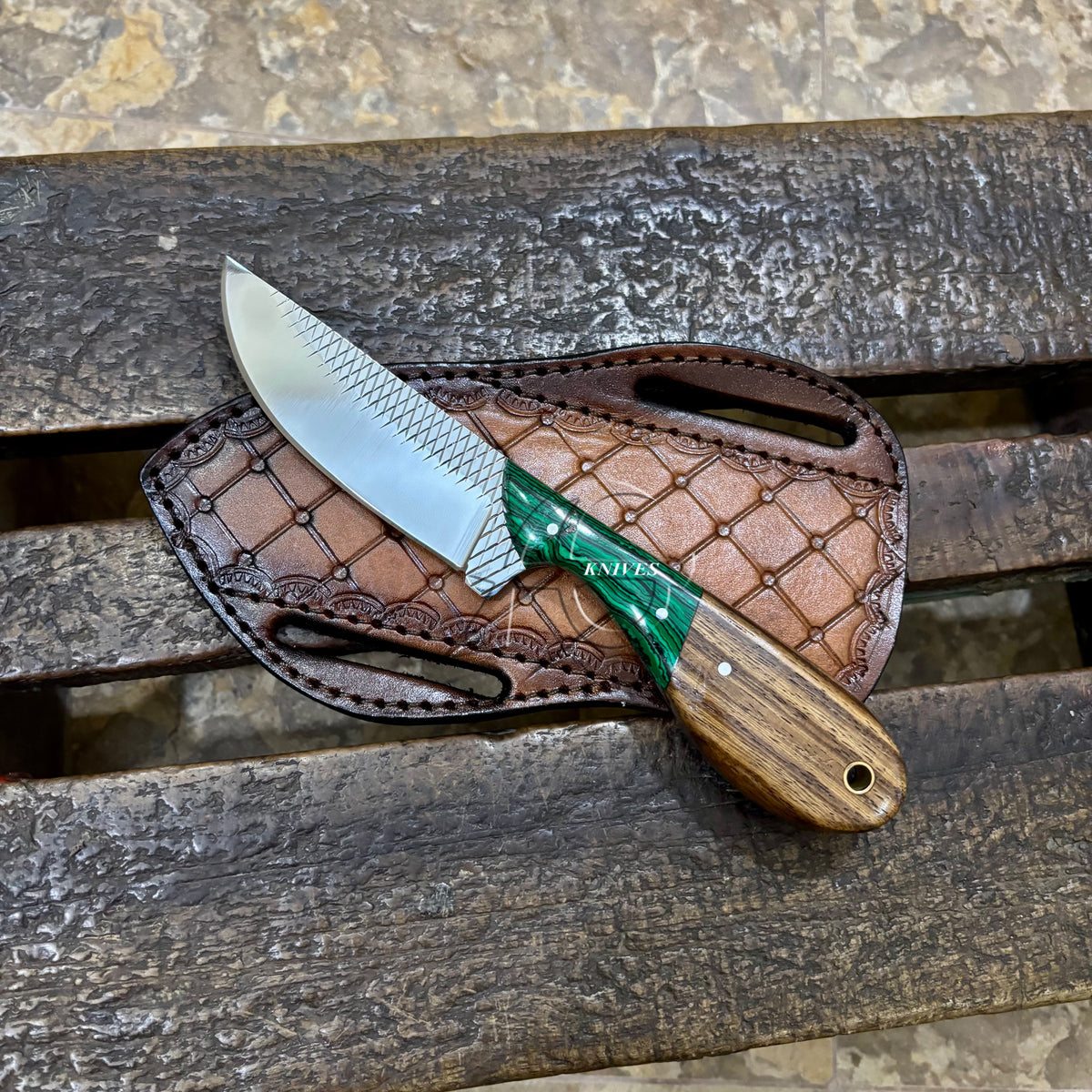 A fixed-blade knife with a textured silver blade, green handle scale, and brown wood-grain handle, resting on a brown leather sheath over a dark wooden slatted surface.