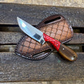 A fixed-blade knife with a silver serrated blade, red and black handle, and brown wooden handle, displayed on a brown leather sheath over a rustic wooden bench.