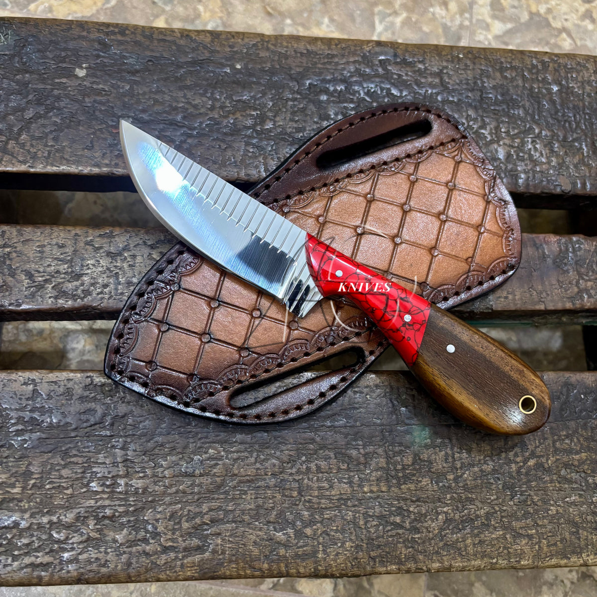 A fixed-blade knife with a silver serrated blade, red and black handle, and brown wooden handle, displayed on a brown leather sheath over a rustic wooden bench.