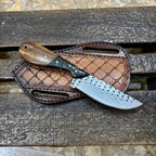 A fixed-blade knife with a dark wood handle and a patterned silver blade rests on a brown leather sheath over a weathered wooden surface.