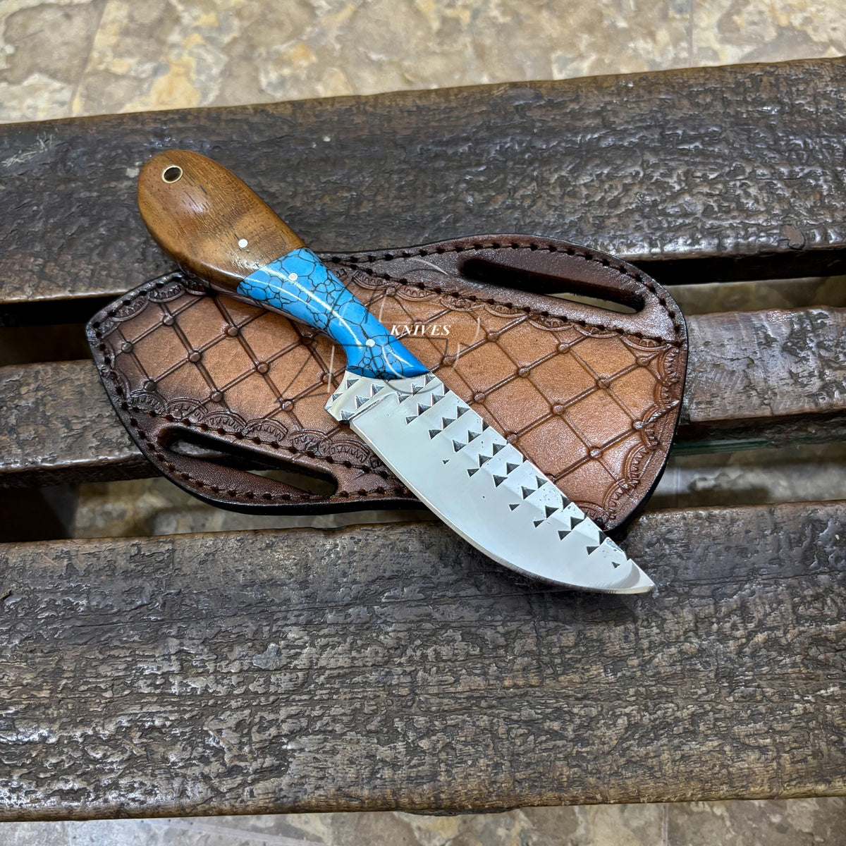 A fixed-blade knife with a brown wooden handle, blue inlay, and a silver serrated blade, resting on a brown leather sheath over a weathered wooden surface.