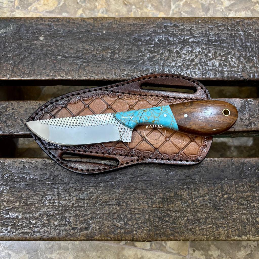 A fixed-blade knife with a silver blade, turquoise handle scale, and brown wooden handle, resting on a brown leather sheath over a rustic wooden surface.