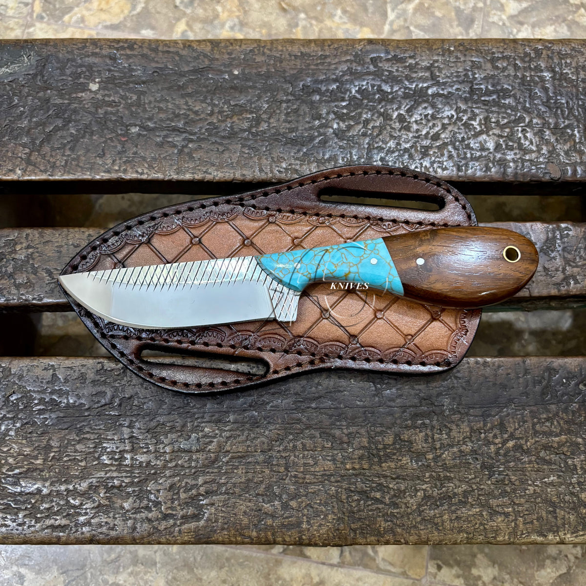 A fixed-blade knife with a silver blade, turquoise handle scale, and brown wooden handle, resting on a brown leather sheath over a rustic wooden surface.