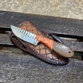 A fixed-blade knife with a patterned silver blade, orange and brown handle, and a brown crocodile-embossed sheath, displayed on a rustic wooden bench.
