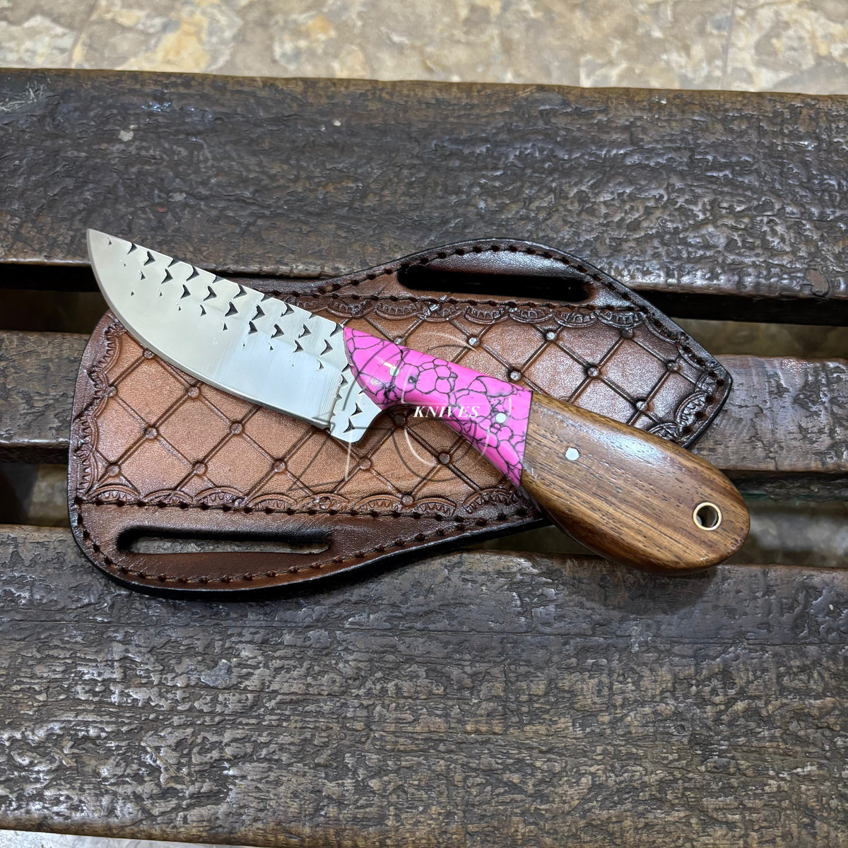 A fixed-blade knife with a patterned silver blade, a bright pink wrapped handle section, and a brown wooden handle, resting on a brown leather sheath over a dark wooden surface.