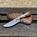 A fixed-blade knife with a silver patterned blade and a brown wooden handle, resting on a brown leather sheath over a weathered wooden surface.