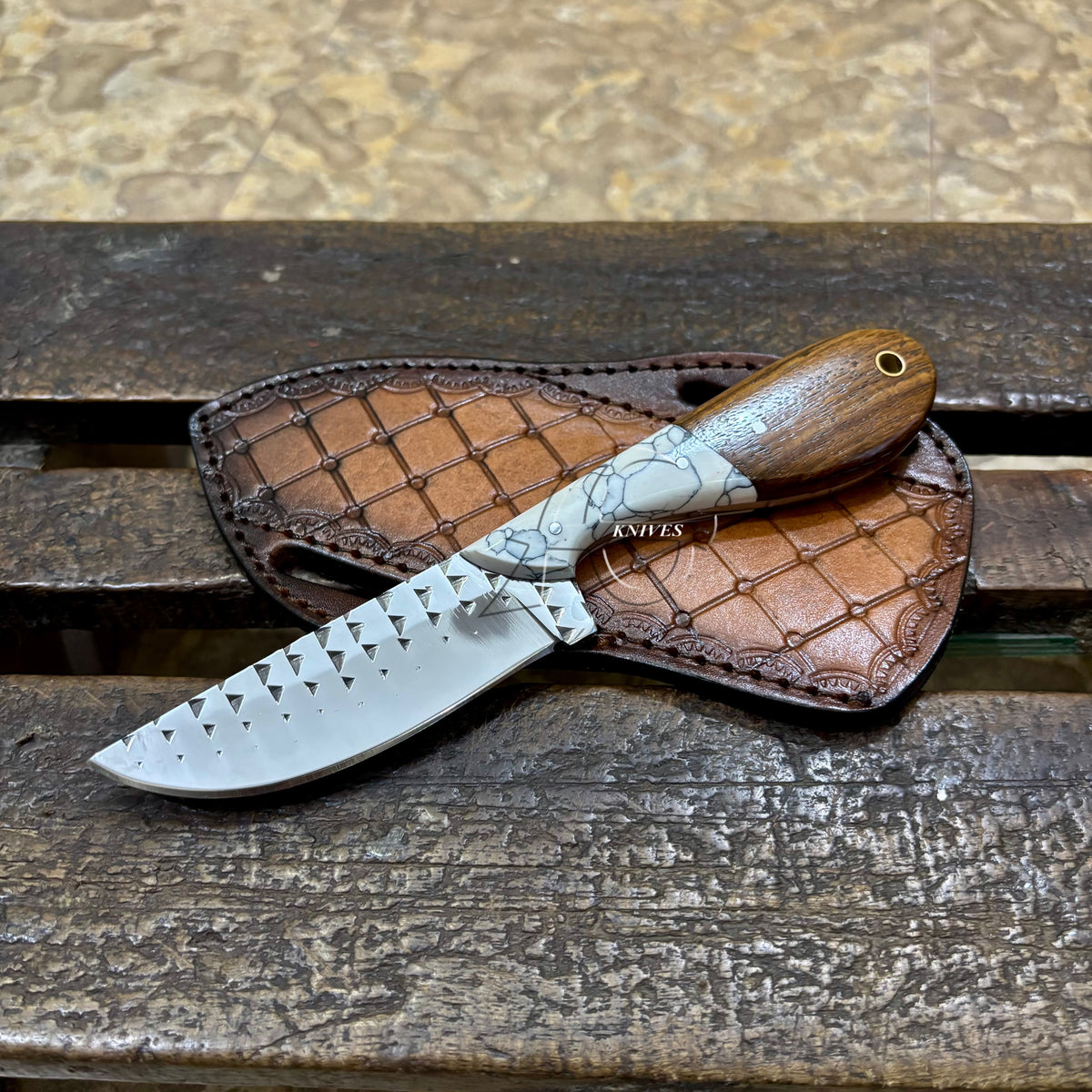A fixed-blade knife with a silver patterned blade and a brown wooden handle, resting on a brown leather sheath over a weathered wooden surface.