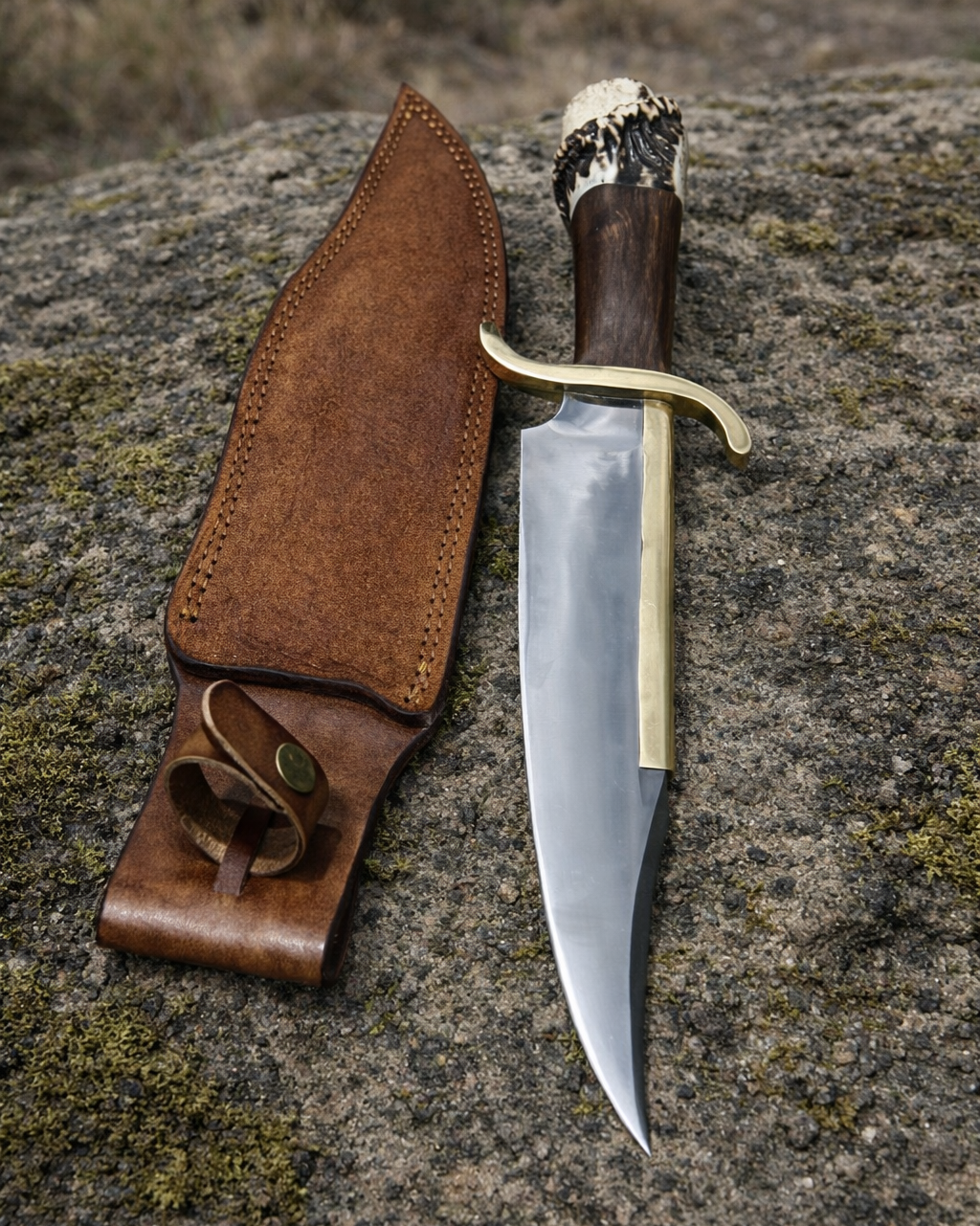 A fixed-blade knife with a polished steel blade, brass guard, dark wood handle, and antler pommel lies beside a brown leather sheath on a large rock outdoors.