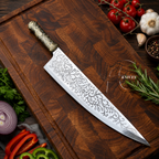 A large chef's knife with an ornate engraved silver blade and a decorative gold-toned handle, placed diagonally on a dark wooden cutting board surrounded by fresh vegetables including tomatoes, garlic, onion, peppers, and rosemary. A watermark reading 'KNIVES' is visible on the blade.