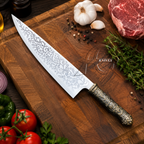 A decorative chef's knife with an etched floral blade pattern and an ornate dark handle, displayed diagonally on a wooden cutting board surrounded by fresh ingredients including tomatoes, garlic, herbs, and a piece of raw meat.