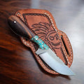 A fixed-blade knife with a dark wood handle and turquoise inlay rests on a brown leather sheath featuring a carved skull design, placed on a dark wooden tabletop.