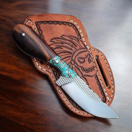 A fixed-blade knife with a dark wood handle and turquoise inlay rests on a brown leather sheath featuring a carved skull design, placed on a dark wooden tabletop.