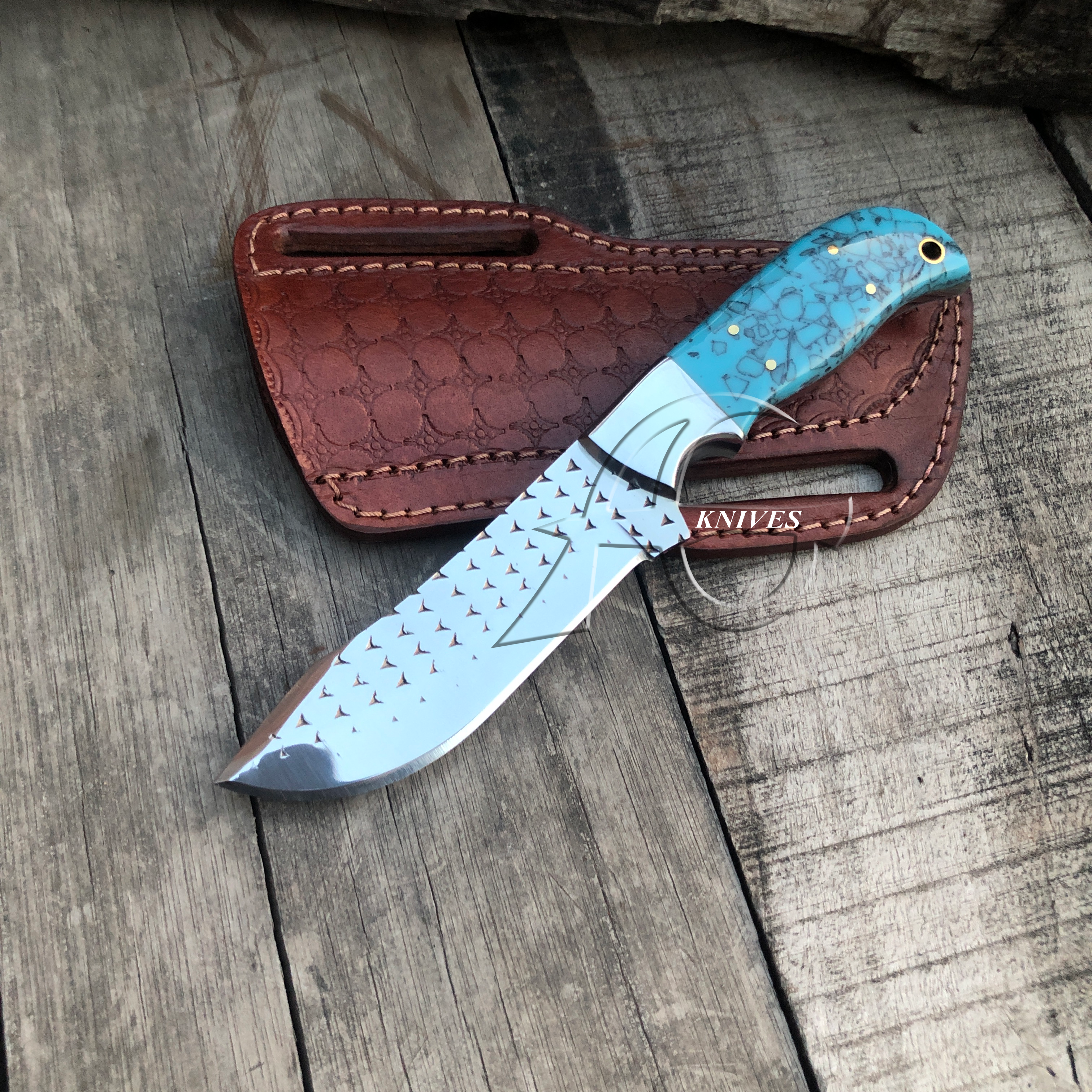 A custom fixed-blade knife with a textured turquoise handle and a patterned silver blade, displayed diagonally on its brown textured leather sheath over a rustic wooden plank background.