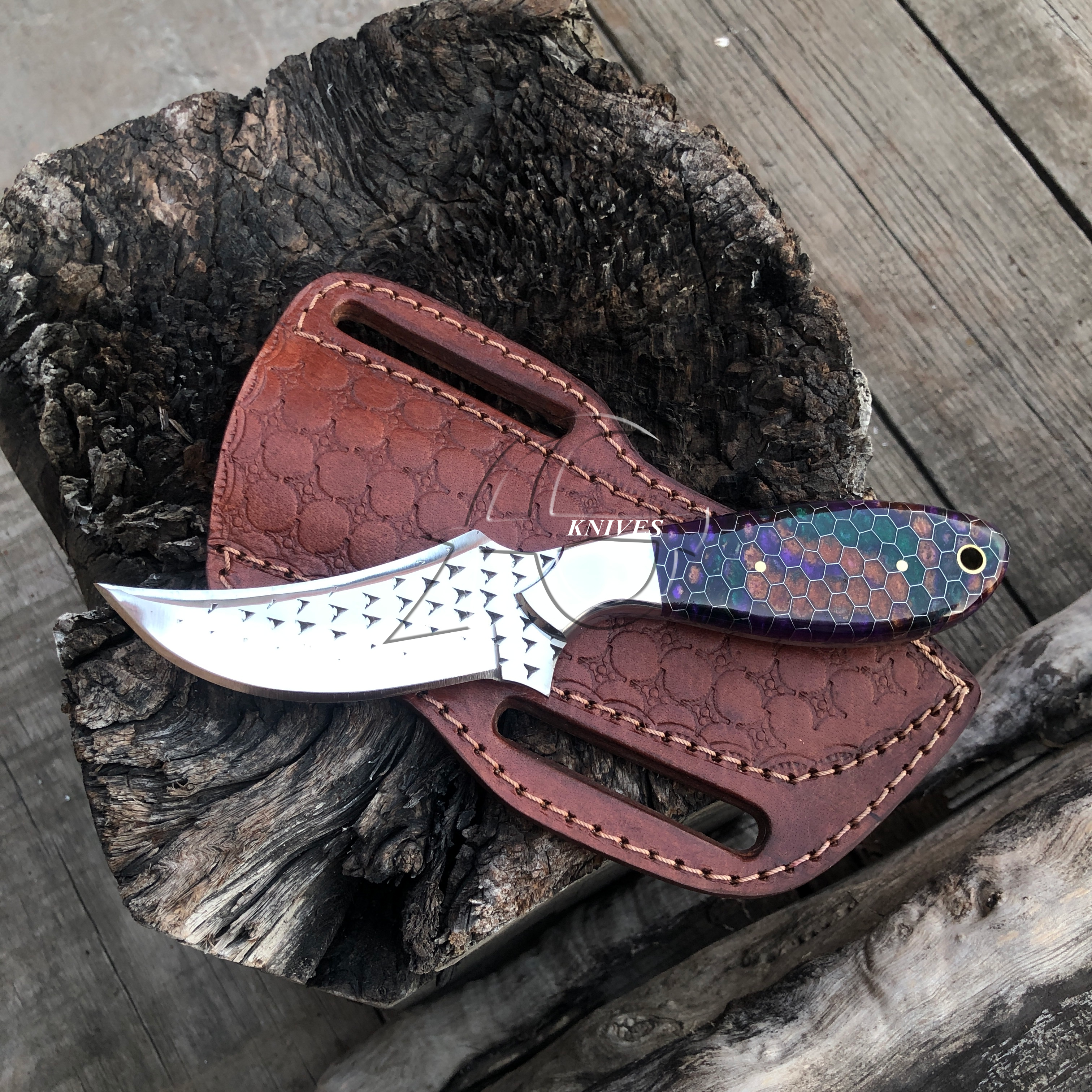 A fixed-blade knife with a rasp file steel blade and a purple-blue honey comb handle scale, resting on a brown embossed leather sheath over a rustic wood log.