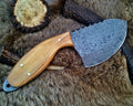 A fixed-blade knife with a Damascus steel blade and a light wood handle, displayed on a brown fur background with antler and wood slice props.