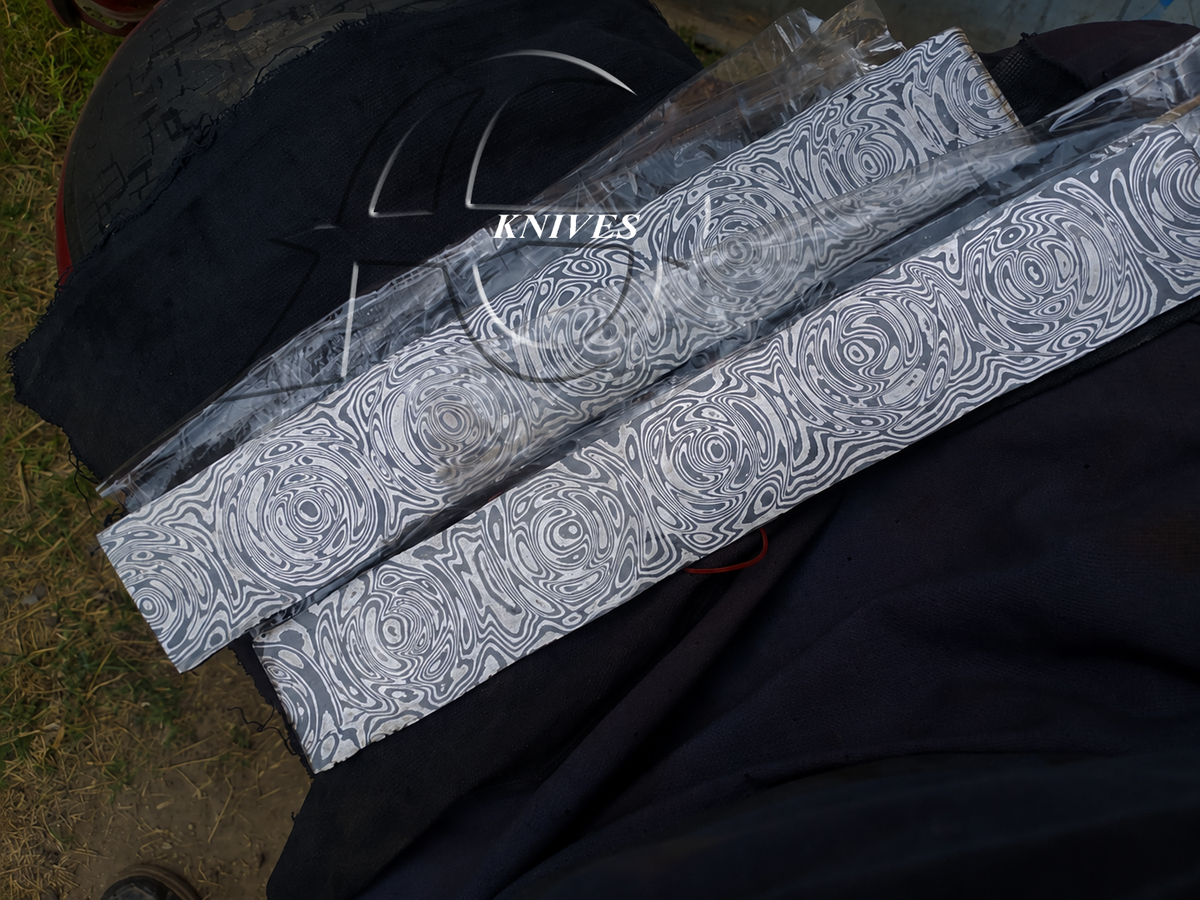 Two long knives with a black and white swirling Damascus-style pattern, wrapped in clear plastic, resting on a black fabric surface outdoors.