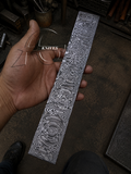 A hand holds a long, narrow Damascus steel knife blank with intricate swirling patterns, set against a dark, cluttered workshop background.
