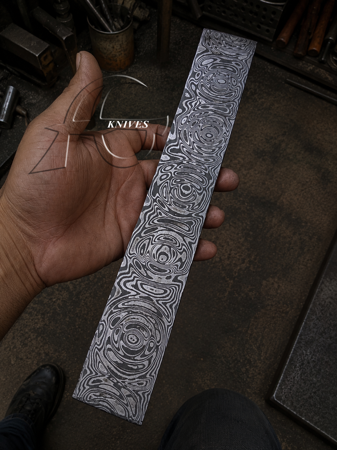 A hand holds a long, narrow Damascus steel knife blank with intricate swirling patterns, set against a dark, cluttered workshop background.