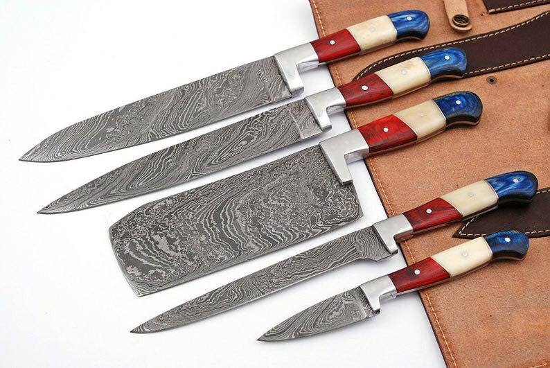 Five Damascus steel kitchen knives with colorful handles in red, cream, and blue, arranged diagonally on a brown leather knife roll against a white background.