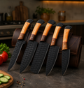 Five kitchen knives with black hammered blades and light wood handles are displayed in front of a brown leather knife roll on a dark countertop, with kitchen props including tomatoes, cucumber slices, and a cutting board in a blurred kitchen background.