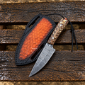 A Damascus steel fixed-blade knife with a patterned wood handle and black cord lanyard, displayed next to its black sheath with a textured orange-red interior, set on a weathered wooden plank surface.