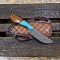 A fixed-blade knife with a patterned steel blade, a brown wooden handle, and a turquoise-colored inlay, displayed on a brown leather sheath over a weathered wooden bench.
