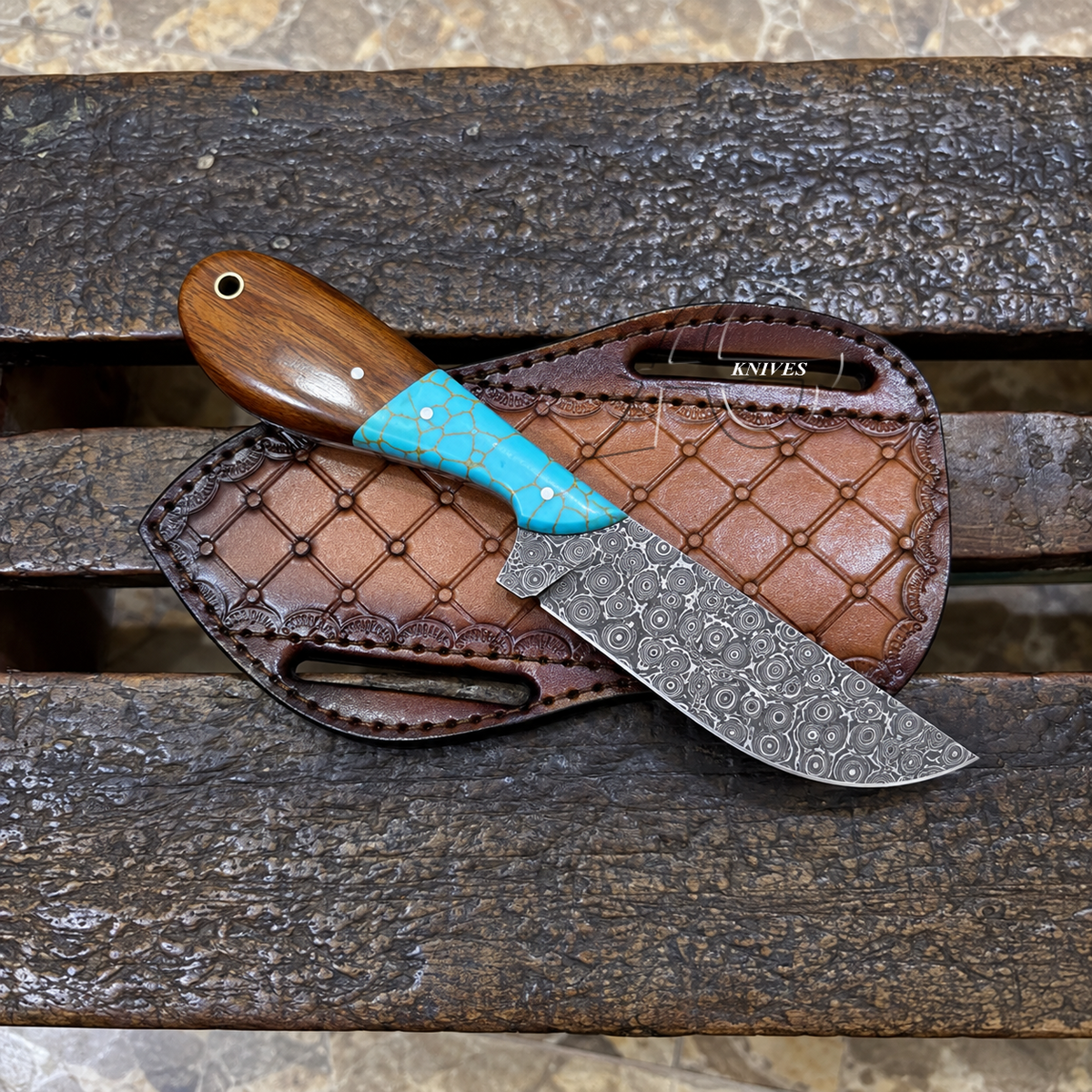 A fixed-blade knife with a patterned steel blade, a brown wooden handle, and a turquoise-colored inlay, displayed on a brown leather sheath over a weathered wooden bench.