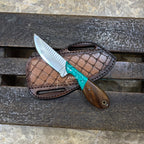 A fixed-blade knife with a silver blade, turquoise wrapped handle, and brown wooden handle, resting on a brown crocodile-embossed leather sheath over a dark wooden plank surface.