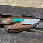 A fixed-blade knife with a brown wooden handle and turquoise inlay rests on a brown leather sheath over a weathered wooden surface.