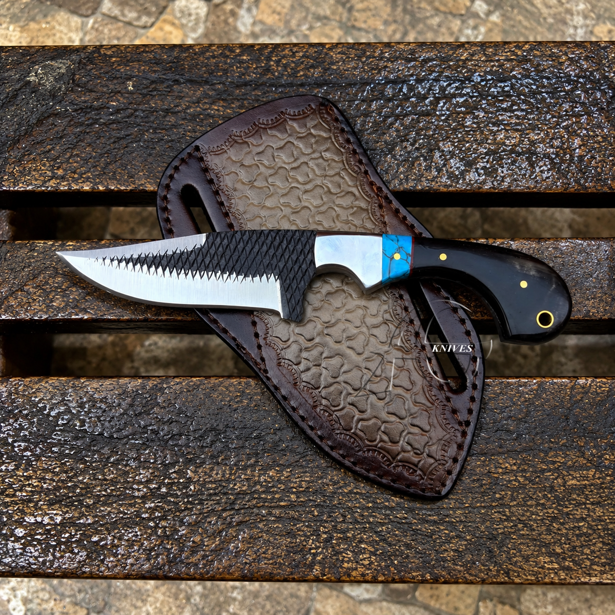 A fixed-blade knife with a partially serrated silver blade, black textured handle, and blue accent near the bolster, resting on a brown embossed sheath over a weathered wooden surface.