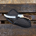 A fixed-blade knife with a serrated silver blade, a cream and brown handle, and a black textured sheath, displayed on a rustic wooden bench.