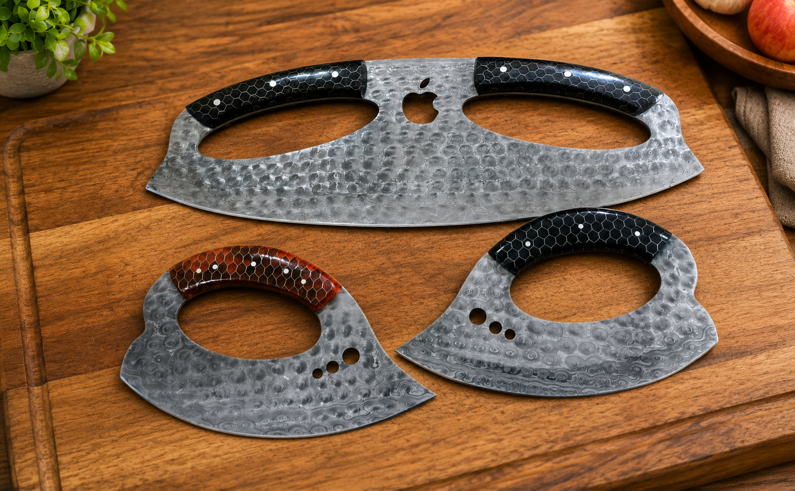 Three handcrafted axe blades with hammered steel finishes and textured black handles, arranged on a wooden cutting board with small decorative plant and fruit props in the corners.