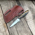 A fixed-blade knife with a silver blade and a blue, gold, and black patterned handle, shown next to a brown leather sheath on a weathered wooden surface. A watermark reading 'KNIVES' is visible across the handle.