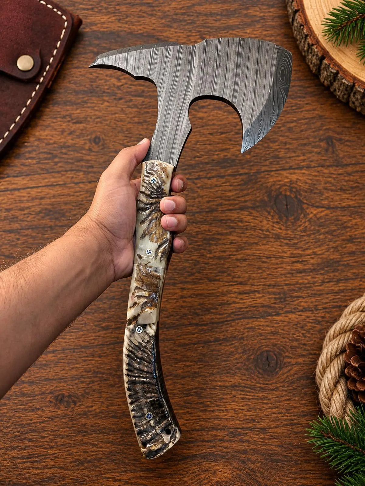 A hand holds a double-bitted axe with a textured gray steel head and a detailed camouflage-patterned wooden handle on a rustic wooden table.