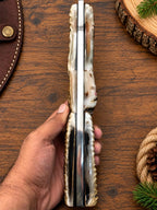 A hand holding a folding knife with a polished bone handle and a silver metal blade, set against a rustic wooden background with a leather sheath and wood slice props.