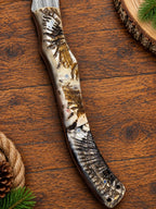 A decorative knife with a highly detailed, realistic animal fur pattern handle, shown on a rustic wooden background with pine needles and a pinecone.