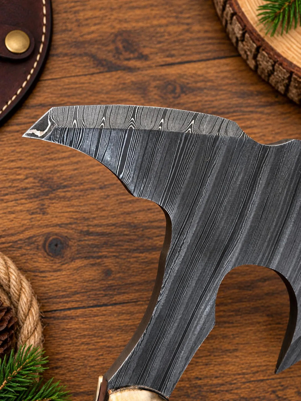 A close-up of a dark forged steel axe head with a textured Damascus pattern, resting on a rustic wooden surface with pine needles and wood slices.