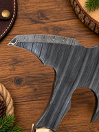A close-up of a dark forged steel axe head with a textured Damascus pattern, resting on a rustic wooden surface with pine needles and wood slices.