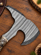 A close-up of a hand axe or hatchet with a textured gray steel head and a carved bone or antler handle, placed on a wooden surface with rustic decorative props.