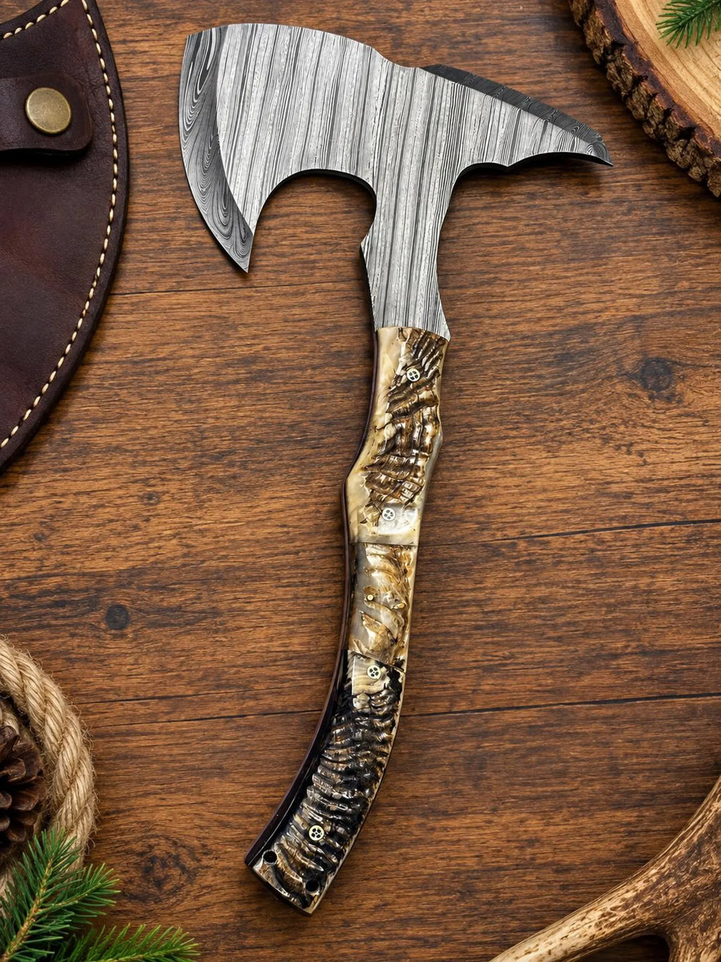 A hand-crafted bearded axe with a textured Damascus steel head and a carved burl wood handle, displayed diagonally on a rustic wooden surface with a leather sheath partially visible.