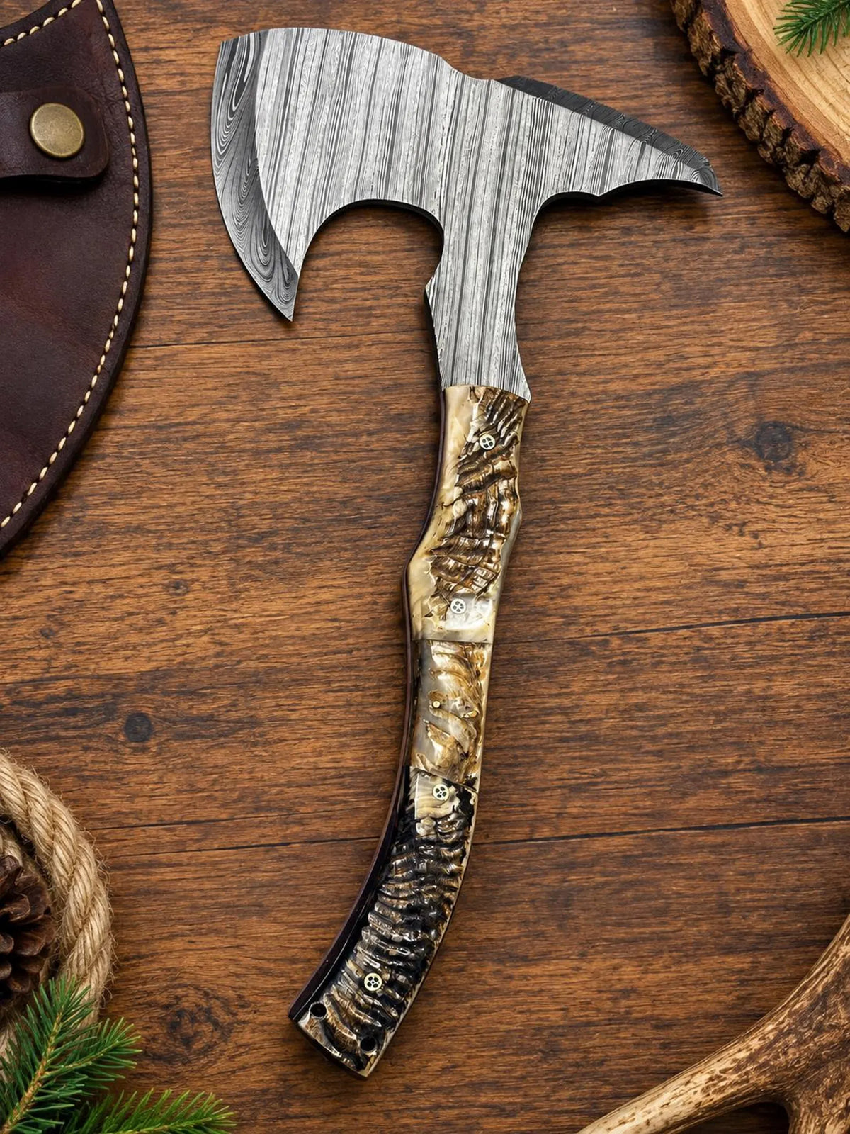 A hand-crafted bearded axe with a textured Damascus steel head and a carved burl wood handle, displayed diagonally on a rustic wooden surface with a leather sheath partially visible.